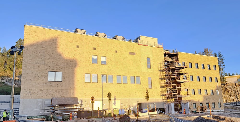 Bilden visar den nya teknikbyggnaden med administrativ byggnad på Hudiksvalls sjukhus. Himlen är blå och molnfri, i förgrunden finns byggarbetsmaterial. Byggnaden är en del av Framtidsbyggets projekt på Hudiksvalls sjukhus. 