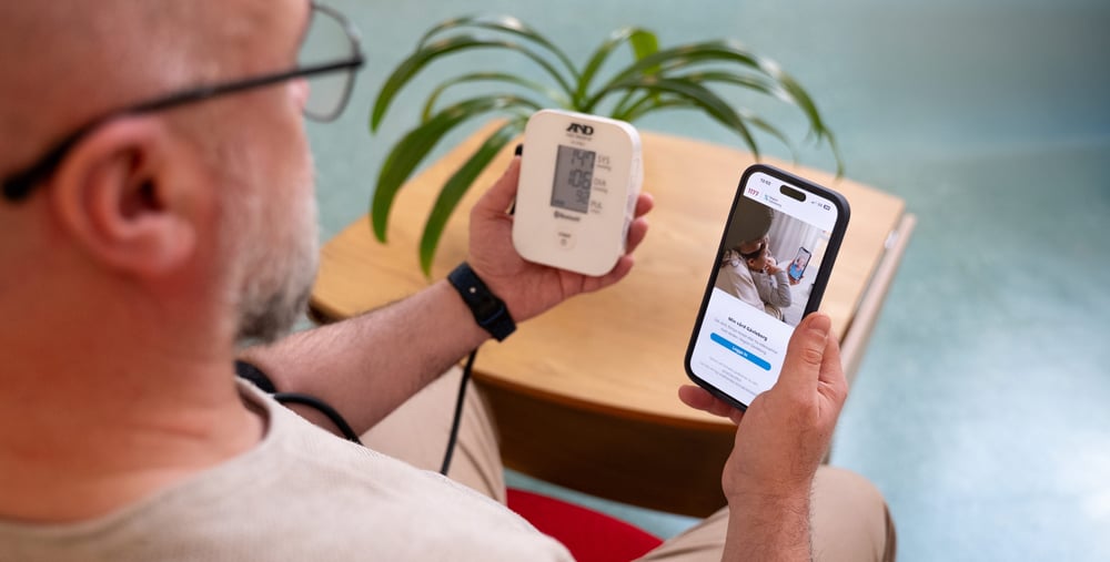 En person sitter ner och mäter sitt blodtryck med en digital blodtrycksmätare. Personen håller mätaren i ena handen och en mobiltelefon i den andra, där en app med vårdrelaterat innehåll visas. 