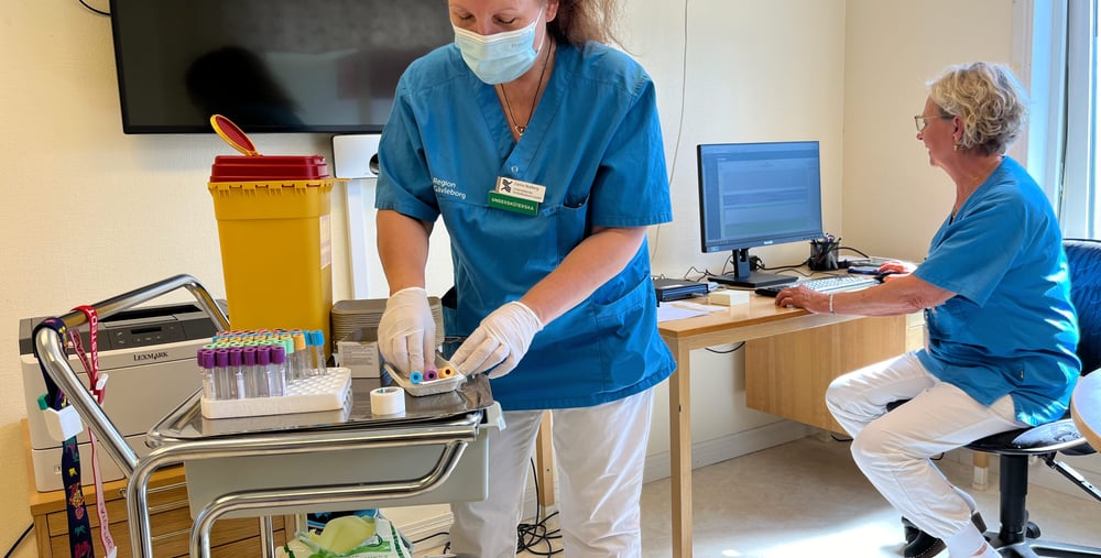 två personer i arbetskläder vid ett rullbord med provrör och vid en dator