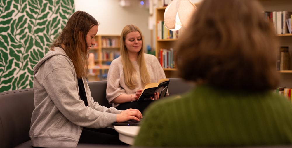Tre personer sitter i en soffa i ett bibliotek; en skriver på laptop, en läser en bok och den tredje är vänd bort från kameran.