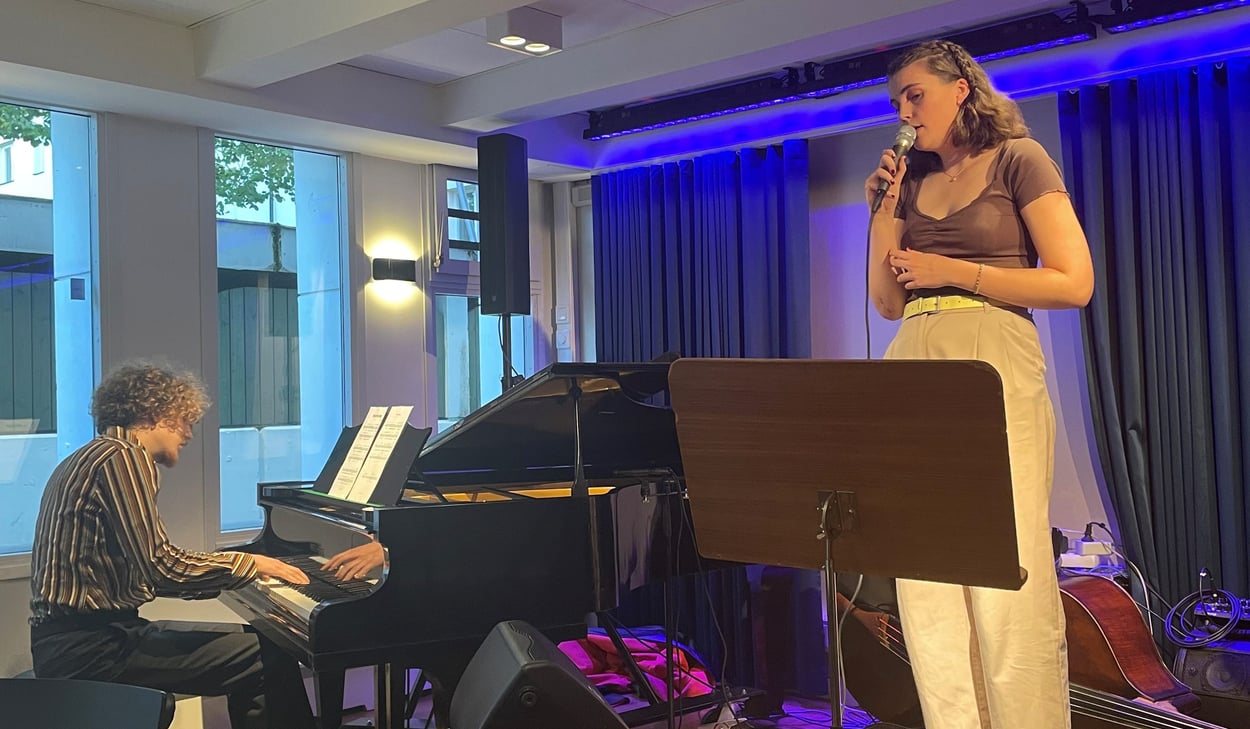 Person står på en liten scen i ett klassrum och  sjunger. En person sitter vid piano och spelar. 