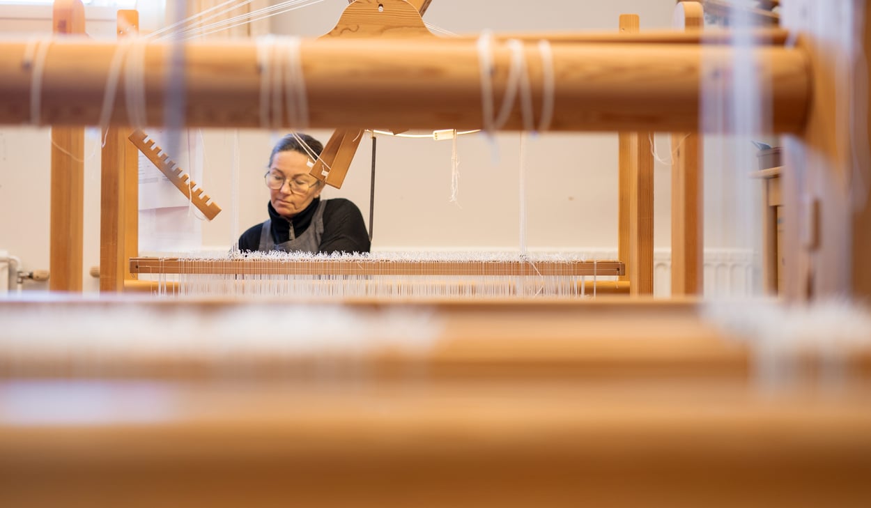 En kvinna syns genom delar av en vävstol medan hon arbetar koncentrerat med vävning.