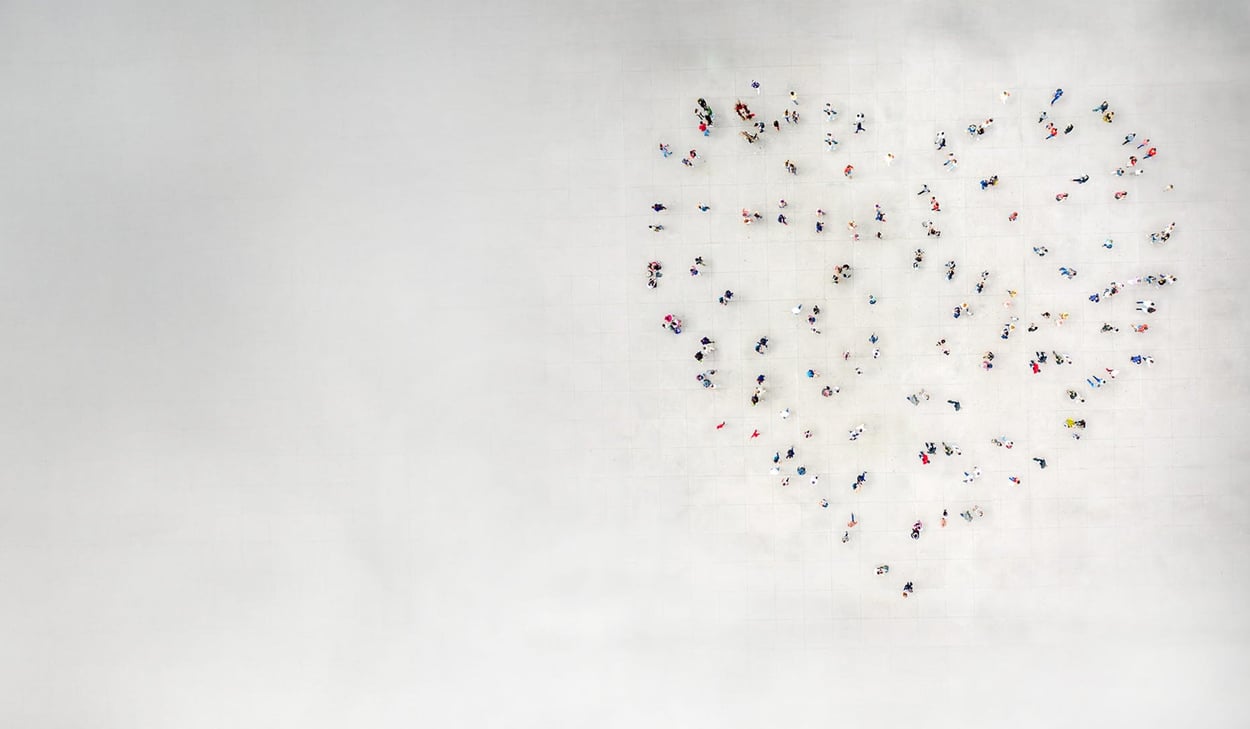 Människor står utspridda så att de tillsammans bildar formen av ett hjärta på en stor öppen yta, sedd uppifrån.