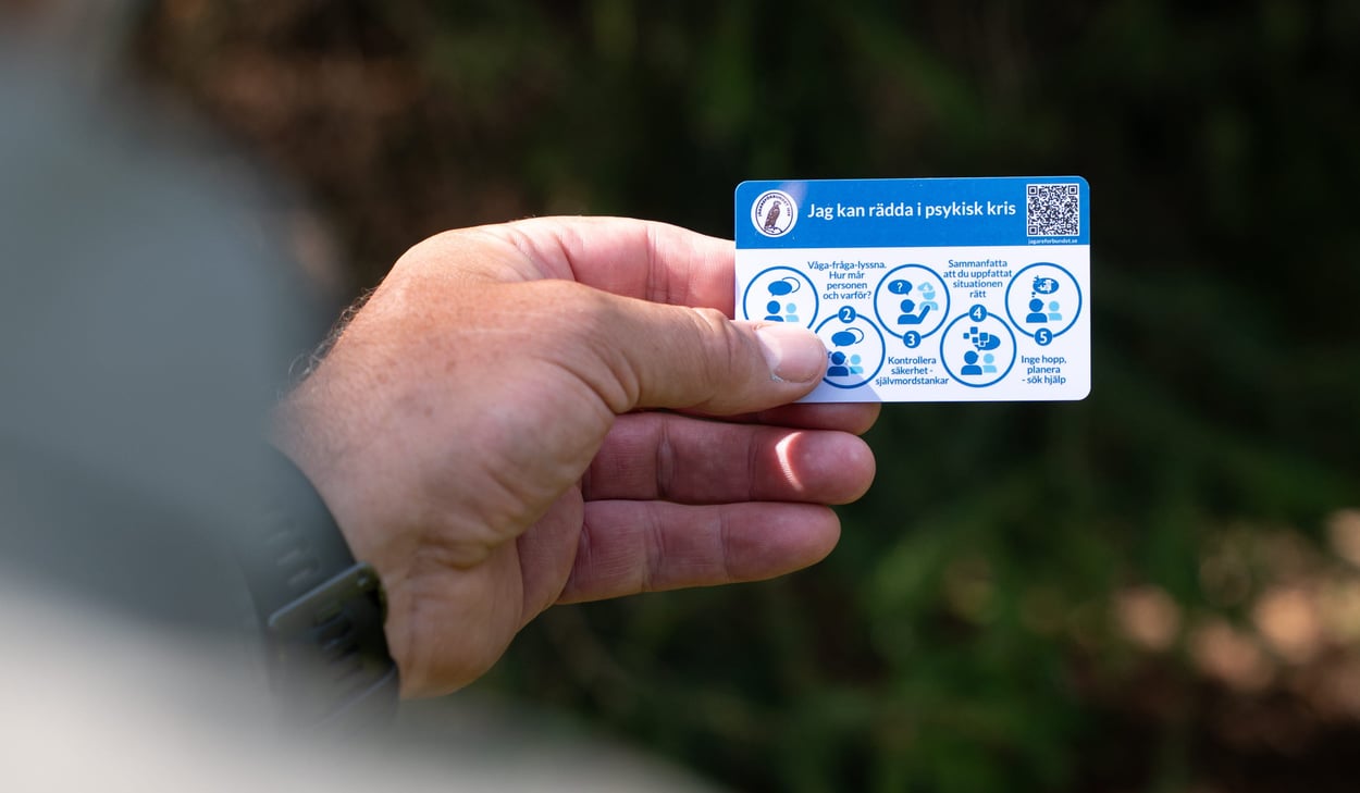En hand håller ett kort med rubriken "Jag kan rädda i psykisk kris" och fem steg för att hjälpa någon i kris, inklusive att våga fråga, kontrollera säkerhet och söka hjälp.