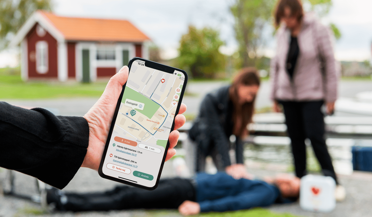 Hand med mobiltelefon, och i telefonen bild på en karta. I bakgrunden en suddig bild på tre personer, där hjärt-lungräddning (HLR) pågår. Bilden är arrangerad och en kvinna står vid sidan, en annan ligger på marken. En tredje ger HLR. 