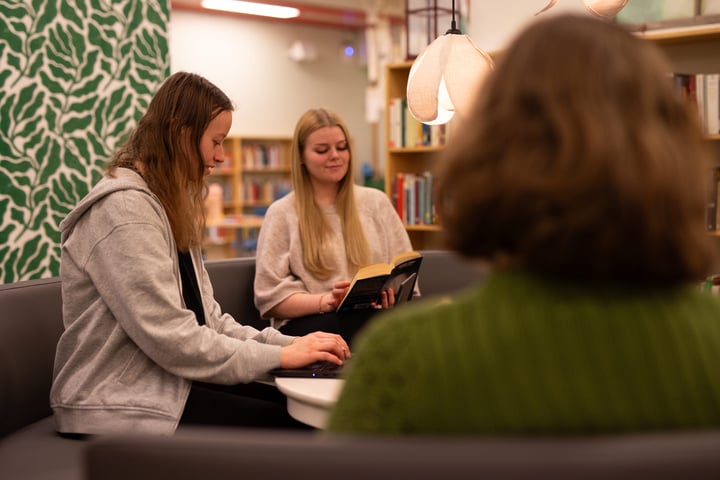 Tre personer sitter i en soffa i ett bibliotek; en skriver på laptop, en läser en bok och den tredje är vänd bort från kameran.